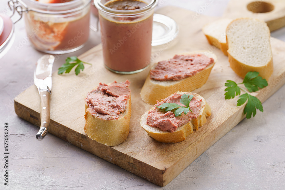Homemade chicken liver pate on fresh french white wheat baguette slices on wooden plate, glass mason jars with cooked liverwurst, top view
