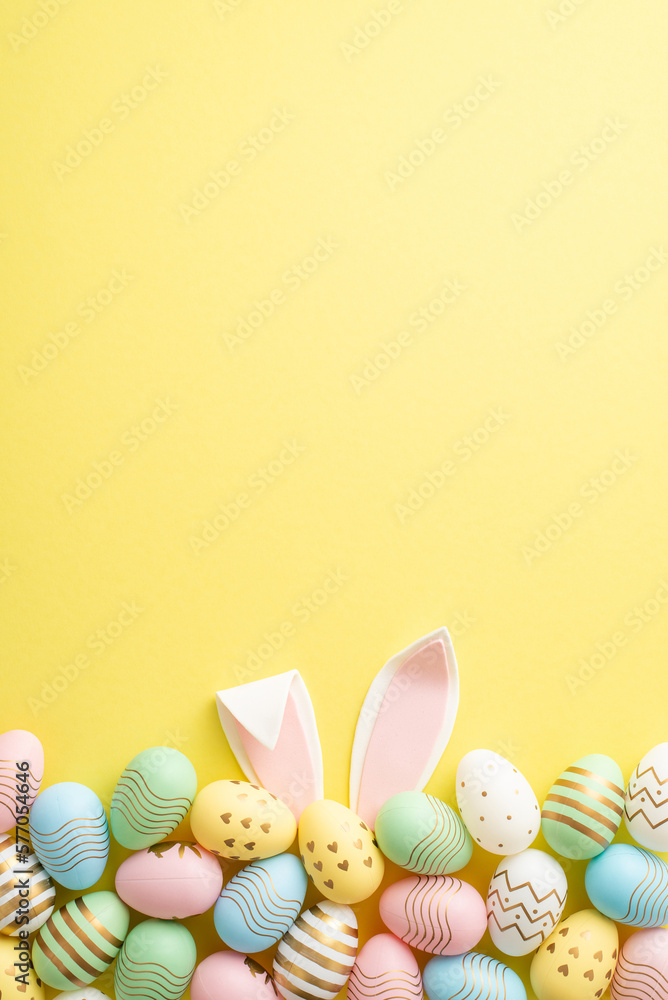 Easter celebration concept. Top view vertical photo of colorful easter eggs and easter bunny ears on isolated yellow background with empty space - obrazy, fototapety, plakaty 