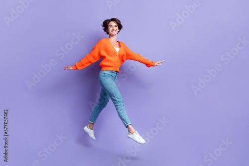 Full length photo of careless good mood youngster active girl orange knitwear denim jeans walking flying pleasure isolated on violet color background