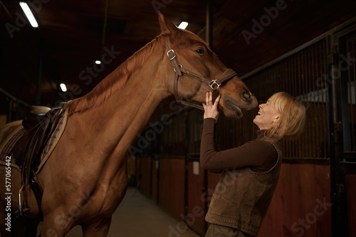 Konstfotografi Happy woman laughing while horse kissing her in stable of riding club