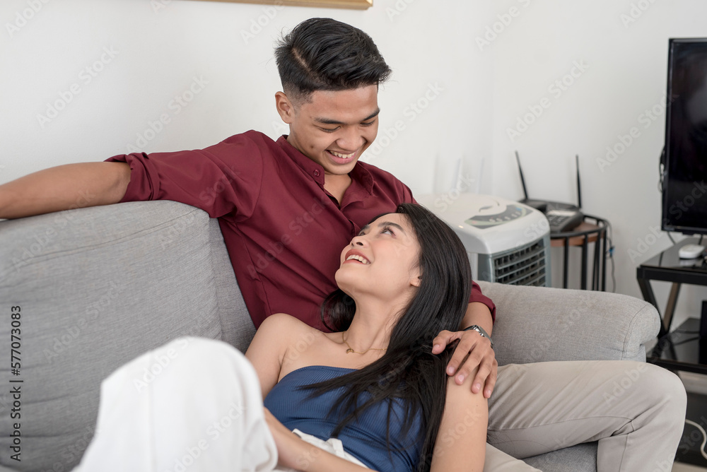 A young asian couple looking fondly, with the woman lying on her partner's lap while sitting at the couch of their apartment. Two people in love.