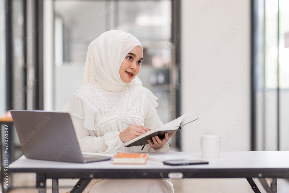 Student muslim woman, Beautiful Asian woman wearing the Islamic headscarf, sitting on a wooden table holding pen writing on notebook and cute smile in workplace with computer. Student muslim woman, Beautiful Asian woman wearing the Islamic headscarf, sitting on a wooden table holding pen writing on notebook and cute smile in workplace with computer.