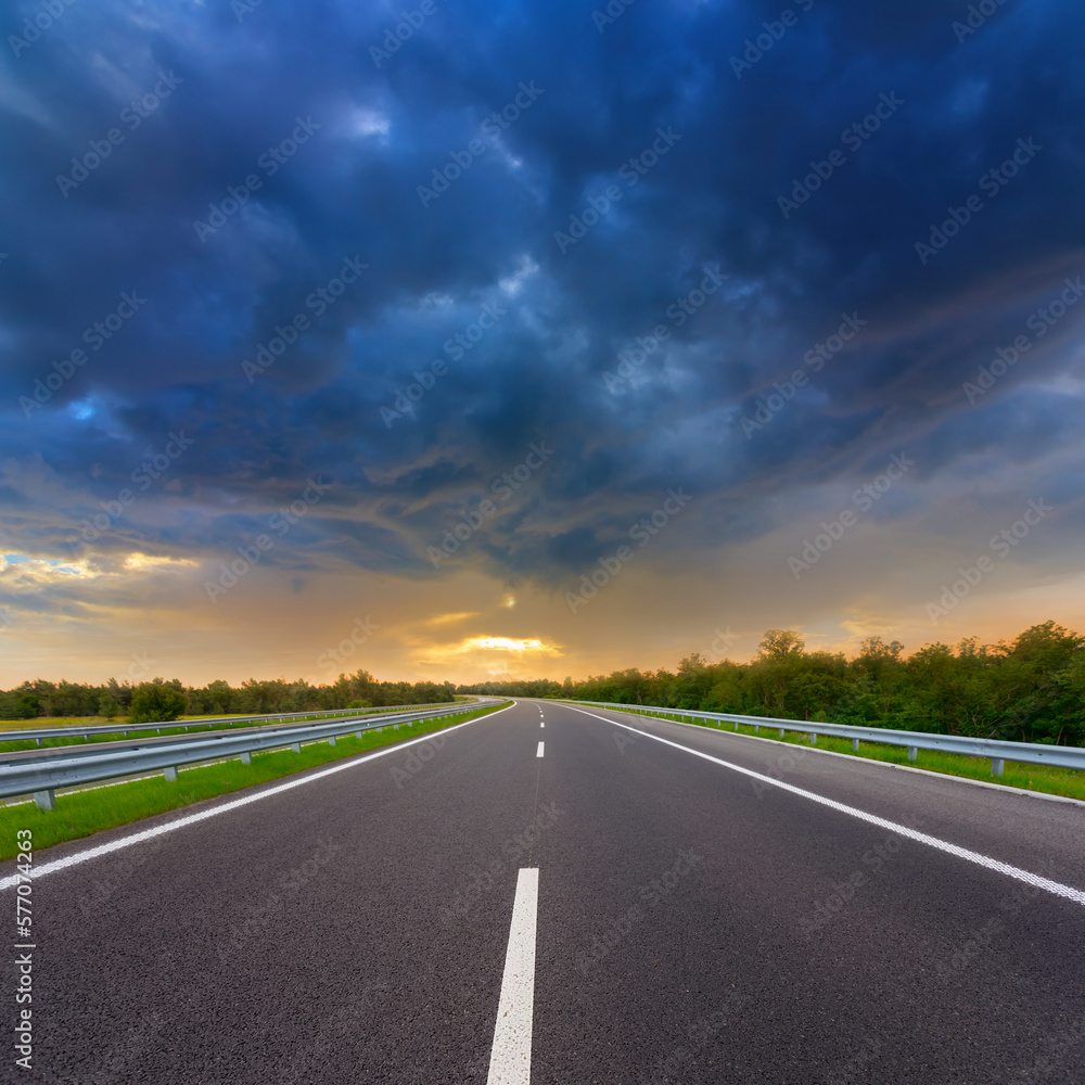 asphalt road leaving far under dense cloudy sky at the sunset, outdoor transportation scene