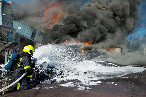 A firefighter uses foam to extinguish a massive plastic waste fire with thick black smoke and flames