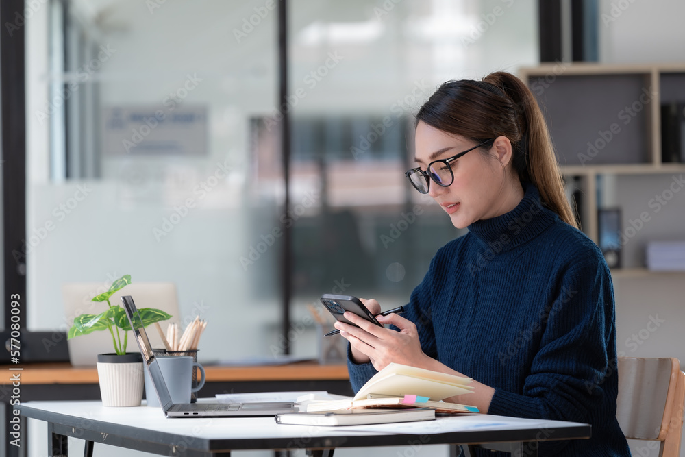 Beautiful Asian woman using a smartphone and working with a laptop while sitting at an office desk, working from home concept. Beautiful Asian woman using a smartphone and working with a laptop while sitting at an office desk, working from home concept.