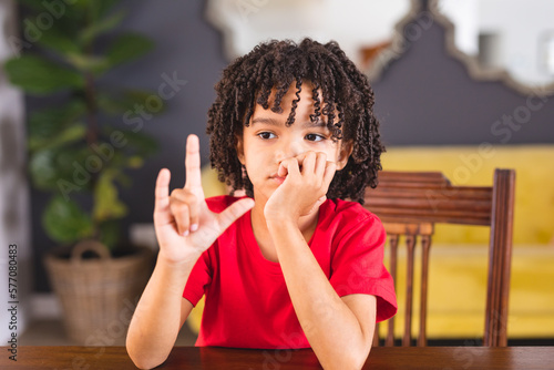 Cute hispanic mute boy showing hand sign while sitting at dining table photo