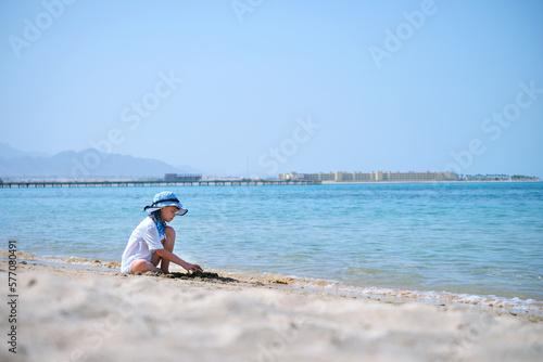 Lonely child girl playing with white sand on sea beach on background of blue sky, clear water and distant shore. Summer tourism and recreation concept