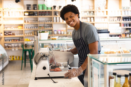 Portrait of smiling young male african american employee using meat slicer at counter in coffee shop photo