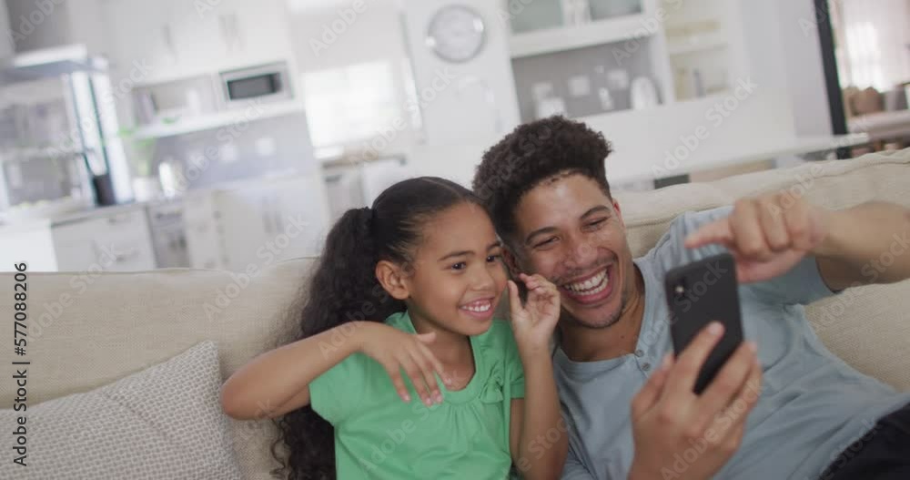 Happy biracial father and daughter sitting on sofa using smartphone Happy biracial father and daughter sitting on sofa using smartphone