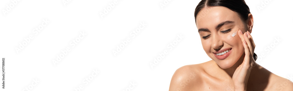satisfied young woman applying moisturizing face cream on cheek isolated on white, banner.