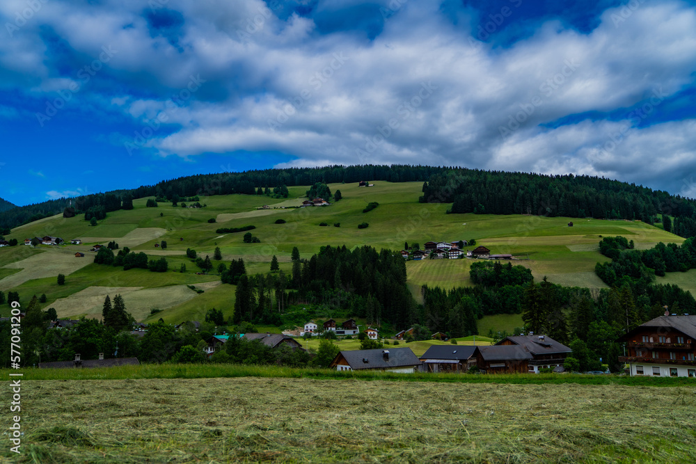 Obraz premium Rural mountain landscape in Italy in the Dolomites. Amazing bright colorful spring and summer landscape. Green blooming fields, wooded mountains and blue sky with clouds. Natural landscape, Europe.