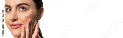 close up view of young woman with face cream on cheek touching face isolated on white, banner.