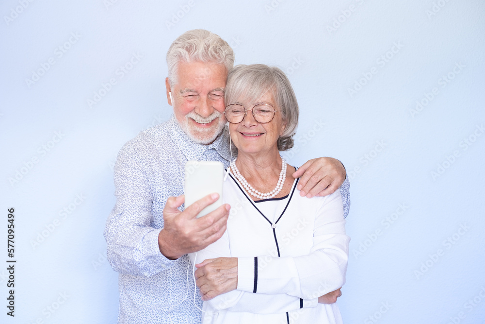 Video call concept. Happy senior couple using smartphone together for video chat online communication, smiling senior people standing on light blue background Video call concept. Happy senior couple using smartphone together for video chat online communication, smiling senior people standing on light blue background