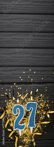 Number 27 blue celebration candle and gold confetti on dark wooden background. 27th birthday card. Anniversary and birthday concept. Vertical banner. photo