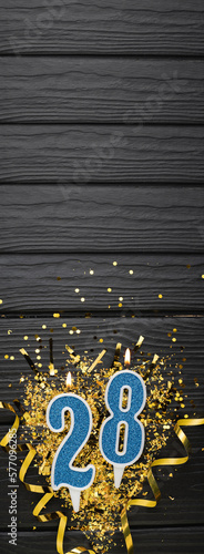 Number 28 blue celebration candle and gold confetti on dark wooden background. 28th birthday card. Anniversary and birthday concept. Vertical banner. photo