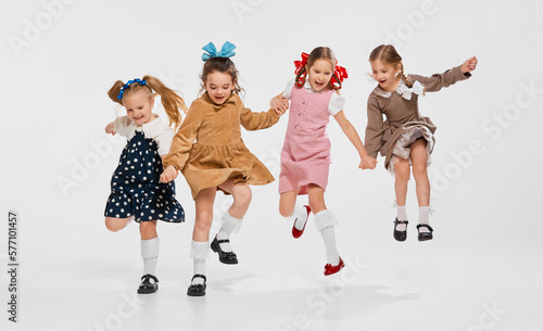 Four beautiful little girls, kids in stylish retro dresses posing, jumping against grey studio background. Concept of childhood, game, friendship, activity, leisure time, retro style, fashion.