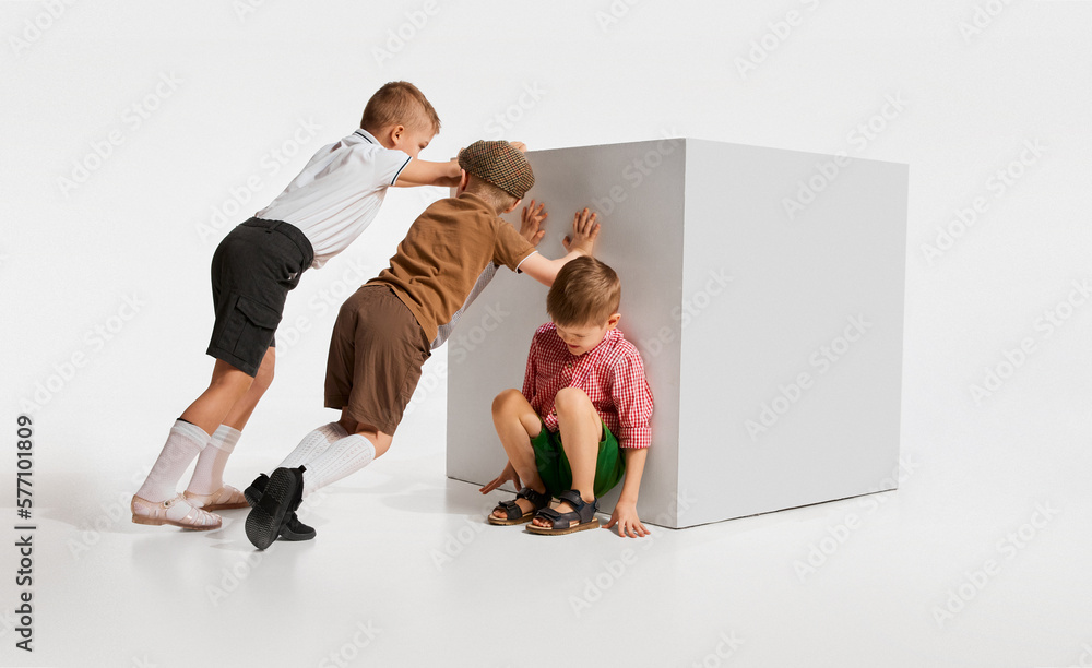 Three little boys kids i retro clothes playing together pushing block forward against grey studio background. Concept of childhood game friendship activity leisure time retro style fashion. Stock Phot...