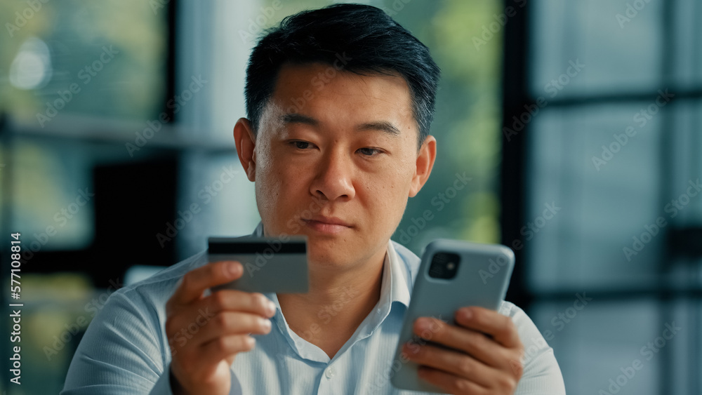 Happy Asian man client buyer businessman use banking mobile phone app at office hold plastic credit card enter secure code buy goods in internet store shopping online cryptocurrency money transaction Happy Asian man client buyer businessman use banking mobile phone app at office hold plastic credit card enter secure code buy goods in internet store shopping online cryptocurrency money transaction
