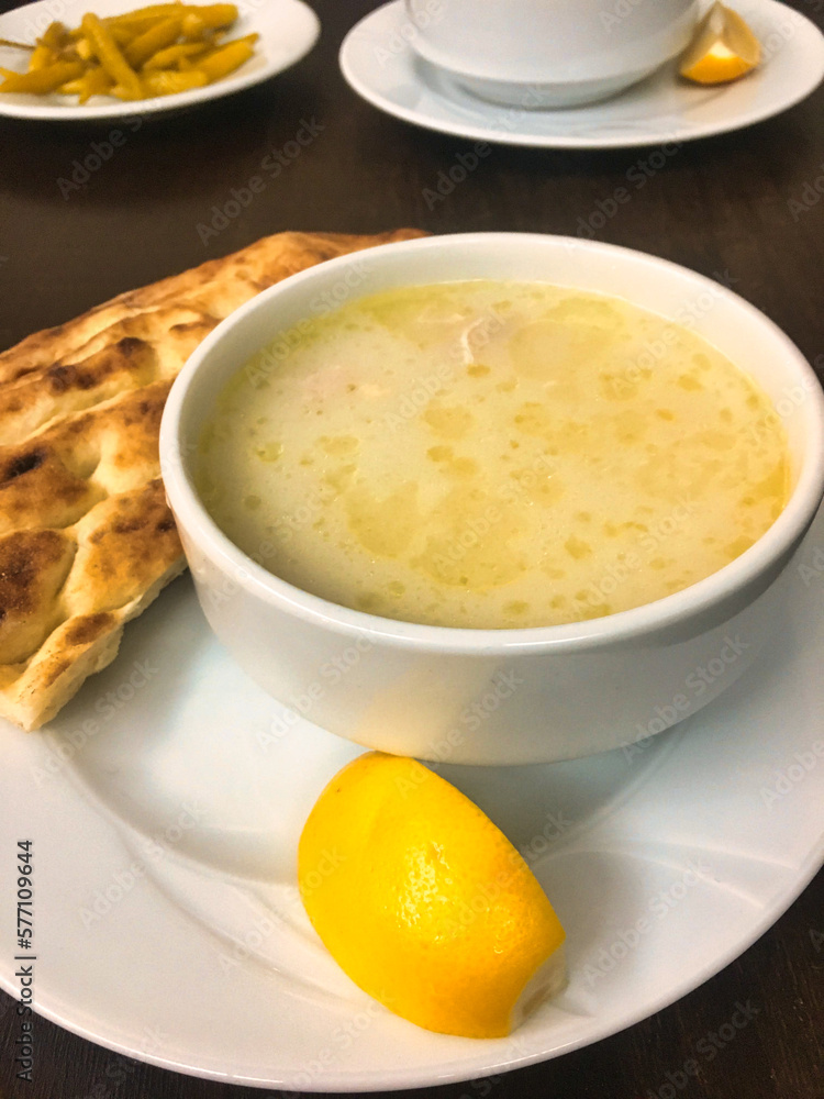 a bowl of chicken broth soup served with a slice of lemon, pickled peppers and pita bread a bowl of chicken broth soup served with a slice of lemon, pickled peppers and pita bread