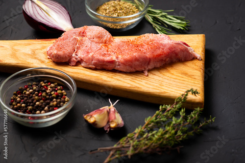 Raw meat steaks on a cutting board. Dark background. Preparation for cooking pork meat. Various spices, seasonings lie nearby.
