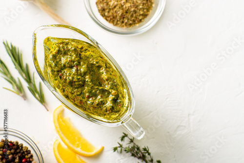 Chimichurri sauce in a gravy bowl on a white background. Various spices lie nearby. Argentinean vegetarian sauce made from olive oil  oregano  parsley. Place for text.