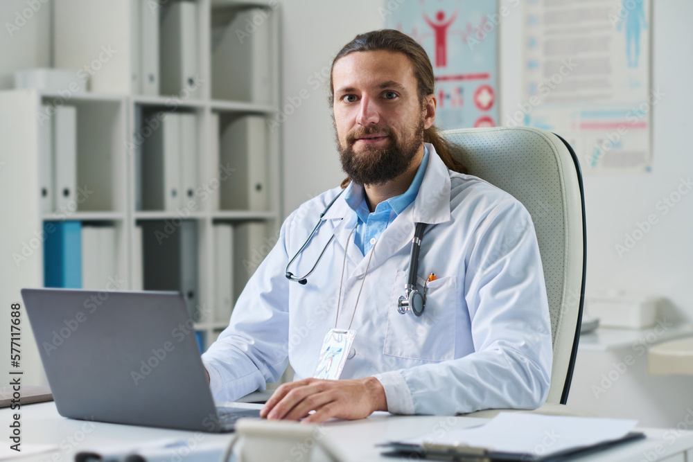 Young confident online doctor in lab coat looking at camera while sitting in front of laptop against shelves with medical documents Young confident online doctor in lab coat looking at camera while sitting in front of laptop against shelves with medical documents