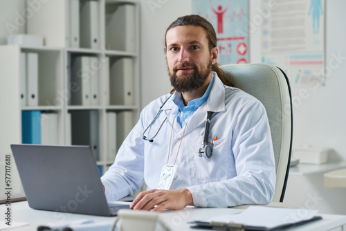 Young confident online doctor in lab coat looking at camera while sitting in front of laptop against shelves with medical documents