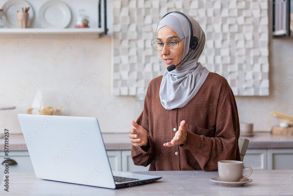 Online training. A young Muslim woman teacher in a hijab, glasses and a headset. He sits at home in the kitchen in front of his laptop and rambles, teaches, teaches via video call. Online training. A young Muslim woman teacher in a hijab, glasses and a headset. He sits at home in the kitchen in front of his laptop and rambles, teaches, teaches via video call.