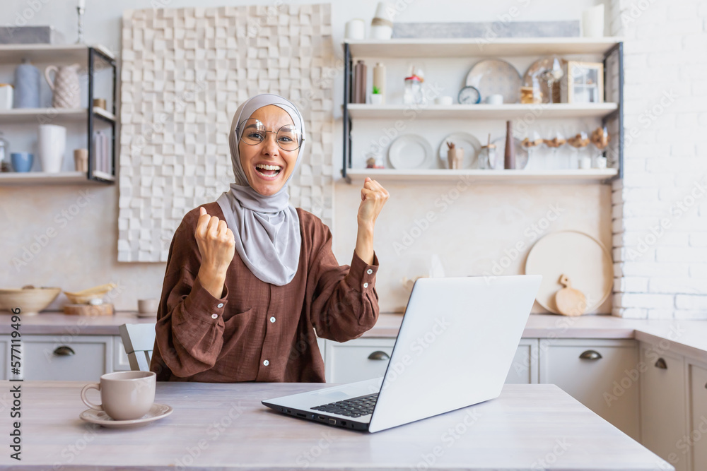 Young happy Muslim student woman in hijab sitting at home, studying on laptop. Shows a yes gesture with his hands to the camera. Passed an online exam, a letter from the university, good results. Young happy Muslim student woman in hijab sitting at home, studying on laptop. Shows a yes gesture with his hands to the camera. Passed an online exam, a letter from the university, good results.