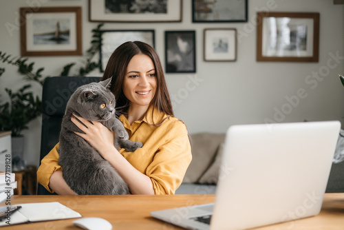 Beautiful young woman smiling on video call via laptop computer, sitting at table at home talking online with family and friends, holding british cat in her arms