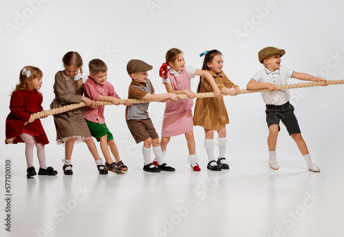 Group of little children, boys and girls playing together, pulling rope against grey studio background. Concept of childhood, game, friendship, activity, leisure time, retro style, fashion.