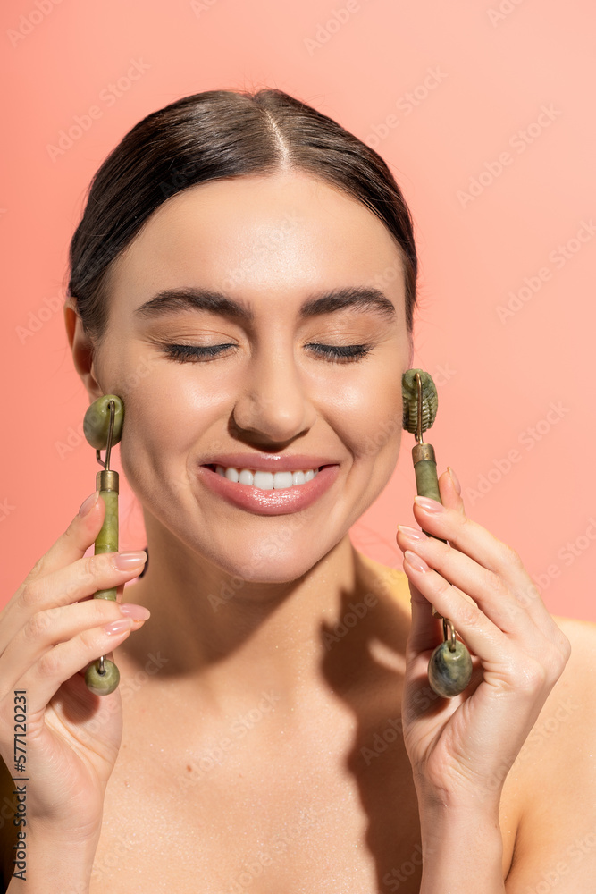 pleased woman doing face massage with jade rollers isolated on pink. pleased woman doing face massage with jade rollers isolated on pink.