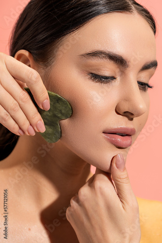 close up view of brunette woman with soft skin doing face massage with jade scraper isolated on pink.