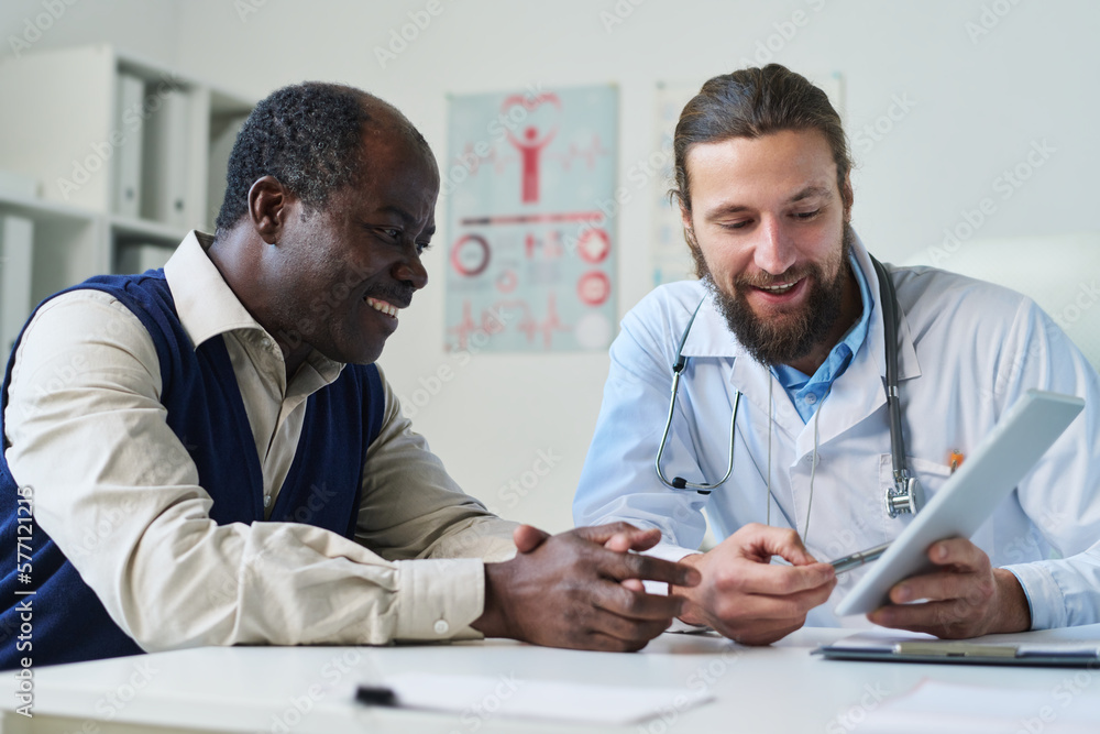 Young smiling doctor pointing at tablet screen with results of medical test and explaining diagnosis to senior African American male patient Young smiling doctor pointing at tablet screen with results of medical test and explaining diagnosis to senior African American male patient