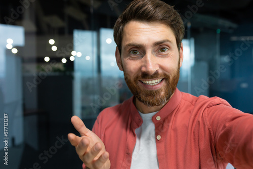 Close-up photo. Young businessman, freelancer, blogger in a red shirt in the office talking on a video call from the phone, smiling at the camera.