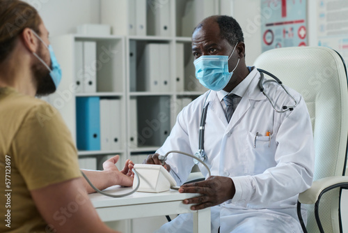 Mature male doctor in protective masks looking at young patient while checking up his blood pressure with tonometer in clinics