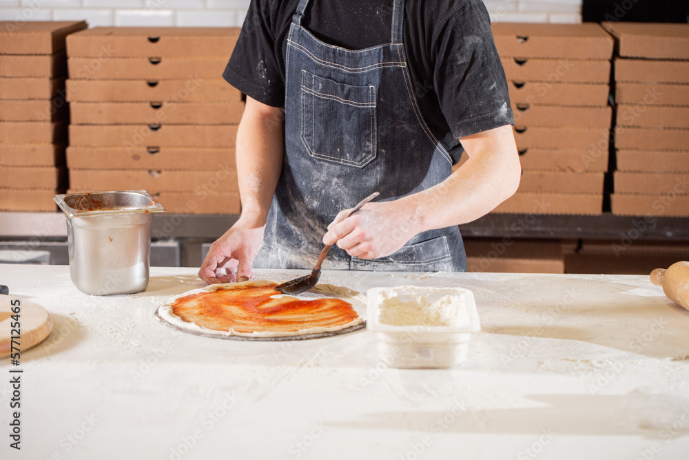 Cooking pizza. the workpiece poured tomato sauce. Closeup hand of chef baker in uniform blue apron cook at kitchen