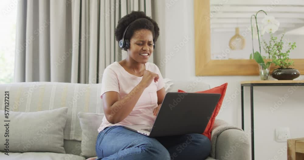 Happy african american woman wearing headphones sitting on sofa, using laptop Happy african american woman wearing headphones sitting on sofa, using laptop
