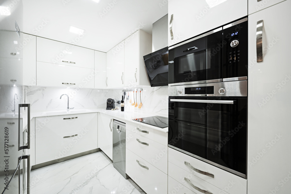 Fototapeta premium kitchen furnished with bright white furniture, smooth white countertops, white marble tiled floors and walls with veins and black glass and stainless steel appliances