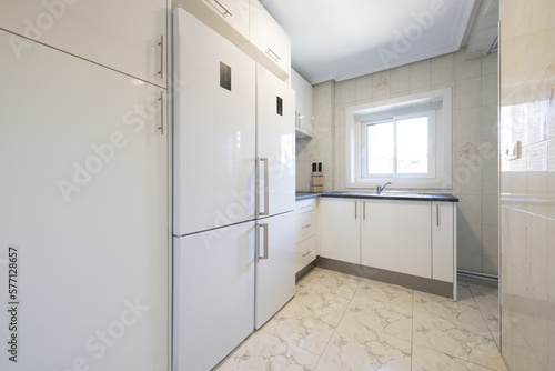 Kitchen furnished with smooth and shiny white cabinets with stainless steel handles  a black countertop and two white attached refrigerators and built-in appliances