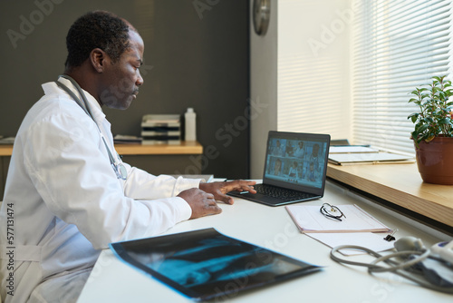 Serious mature African American male radiologist sitting by workplace in front of laptop with group of intercultural doctors on screen