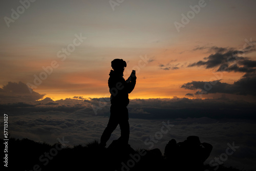 Man taking photos of the sunrise on top of the mountain. Mountaineer is capturing the sunrise with his cell phone. Silhouette of a person. 