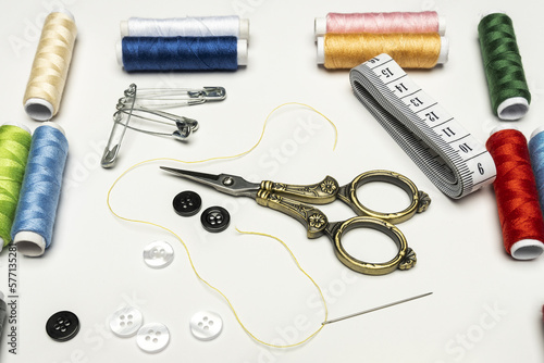 Still life with golden vintage sewing scissors on a white surface along with spools of thread, buttons, needles and thread and a tape measure