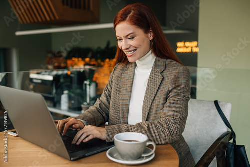 Cheerful woman with ginger hair working in a cafe. Smiling female in formal wear sitting at a table in a coffee shop.