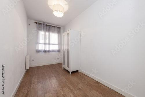 Empty room in an apartment with a matte wooden floor  a small white cabinet and a window with curtains