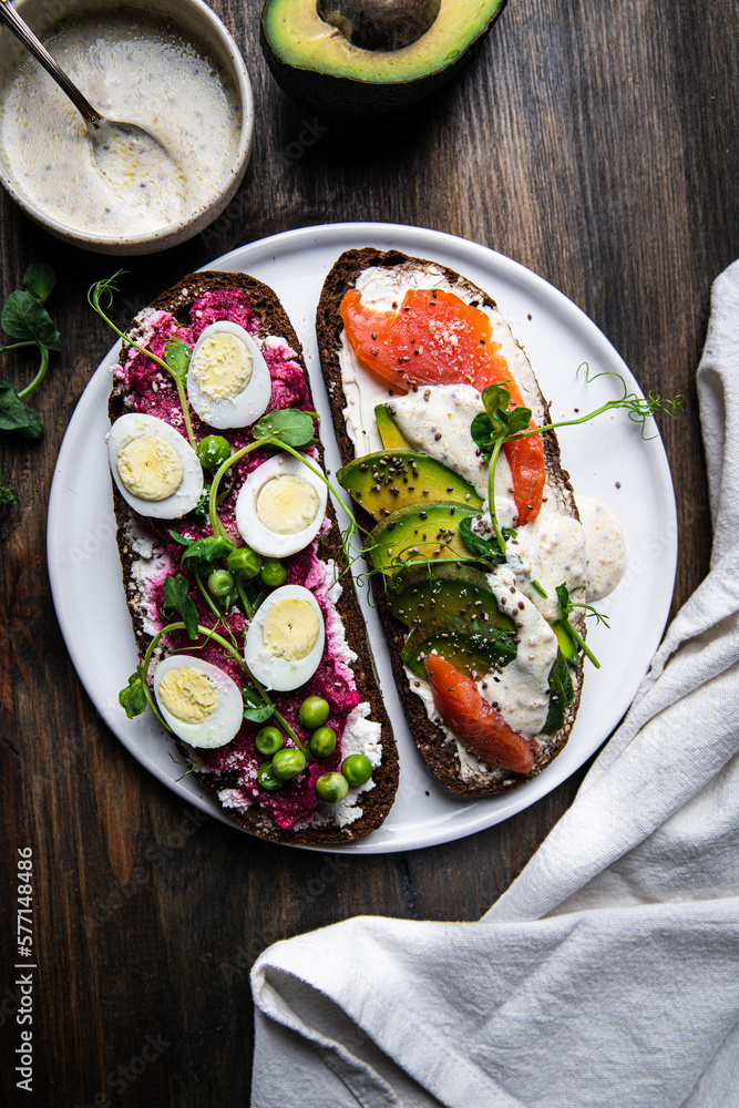 Two smorrebrods (open sandwiches with different toppings: avocado-salmon-cream cheese and quail eggs -beetroot - cream cheese) and mustard sauce on wooden board/