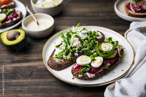 Two smorrebrods (open sandwiches) with different toppings: green peas - cottage cheese and quail eggs -beetroot paste- avokado- cream cheese on rye bread and glass of lemonade on wooden table. 