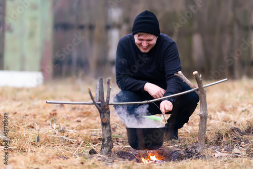Young man with a smile cooks soup on a campfire