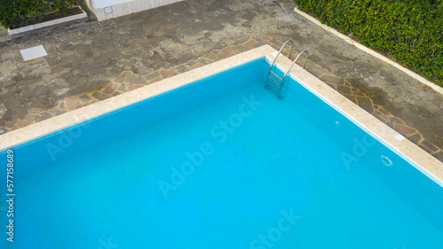 Aerial view of a rectangular swimming pool with ladder  belonging to a large villa. The pool is empty and no one is swimming. Around the water there is a stone floor.