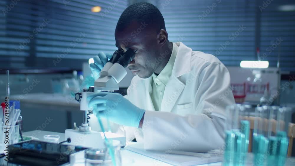 African American scientist in sterile gloves and white coat using microscope and taking notes while doing research in laboratory African American scientist in sterile gloves and white coat using microscope and taking notes while doing research in laboratory